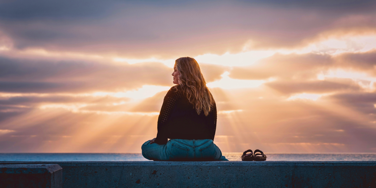 Woman sitting beside the ocean at sunset as rays of sunlight break through clouds over the water.